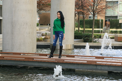 An Indian model on a walkway over a man made pond and concrete columns. She is wearing long, black, straight hair, a green sweater, torn blue jeans and knee high high heels.