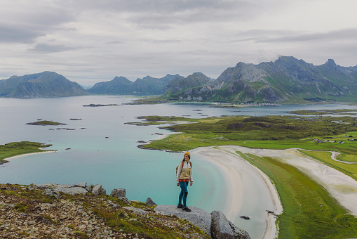 Side view of woman traveler in a yellow hat getting to the top of the mountain, admiring the view of the turquoise-coloured sea, green meadow and dramatic peaks in Northern Norway