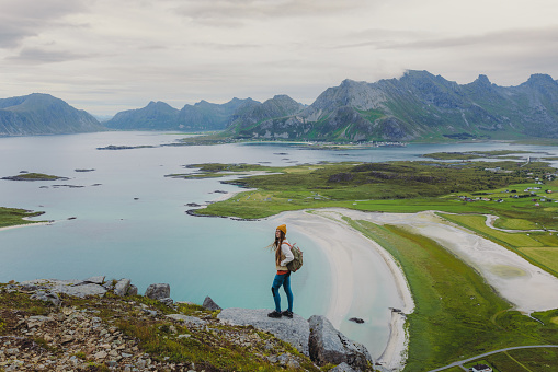 Side view of woman traveler in a yellow hat getting to the top of the mountain, admiring the view of the turquoise-coloured sea, green meadow and dramatic peaks in Northern Norway