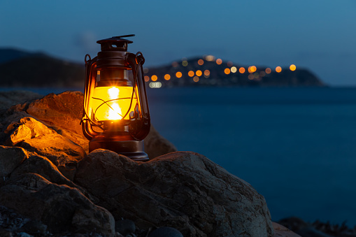 Frontal view of an old oil lantern sitting on a rock of the beach of Cala Regina and city lights bokeh on the background