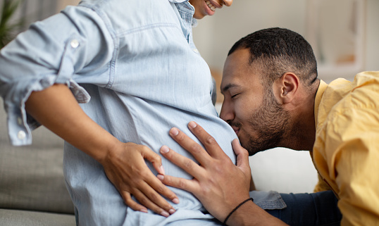 Pregnancy And Childbirth. Cropped Shot Of African American Husband Kissing Wife's Belly Sitting On Sofa At Home. Family Expecting Baby, Love And Happiness. Concept