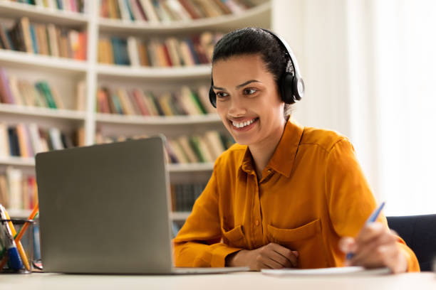 istock Happy mixed race lady having online class, using headset, looking at laptop screen and taking notes, sitting in library 1471477937