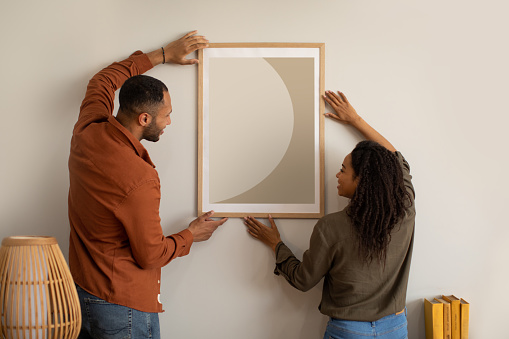 African American Couple Hanging Poster On Wall At Home. Young Spouses Decorating Living Room Together In New House After Renovation. Interior Design Concept