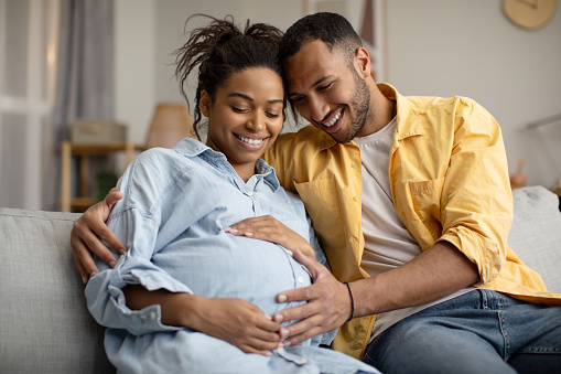 Pregnancy. Black Husband And Pregnant Wife Hugging Sitting At Home. Family Couple Expecting Baby, Man Embracing Woman Smiling Posing Together. Childbirth Concept