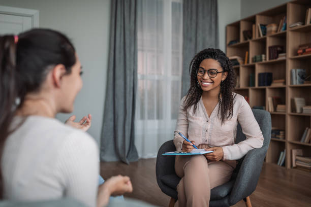 sonriente joven negra doctora psicóloga consultando a una mujer europea en el interior de una clínica moderna - terapia fotografías e imágenes de stock