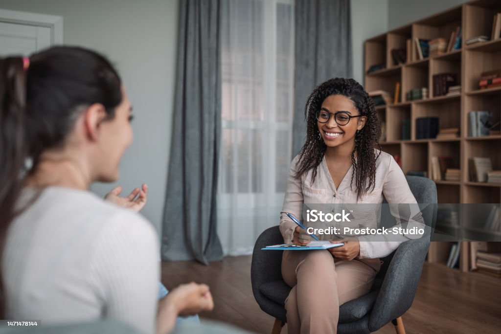 Smiling young black female doctor psychologist consulting european woman in modern clinic interior Smiling young black female doctor psychologist consulting european woman in modern clinic interior. Mental health care, medical support, psychological problems, therapy and professional treatment Mental Health Professional Stock Photo