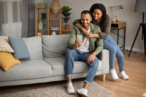 African American Husband And Wife Using Phone Browsing Internet Sitting On Sofa At Home On Weekend. Couple Using Mobile Application On Smartphone. Online Technology, Gadgets