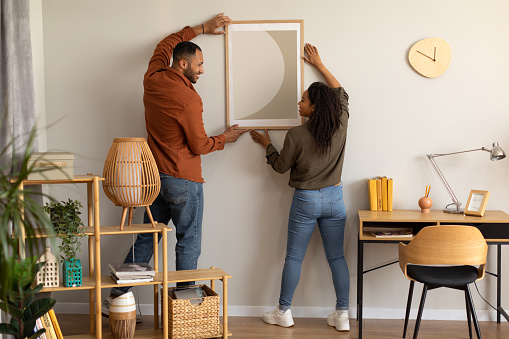 Black Millennial Spouses Hanging Poster On Wall Together At Home