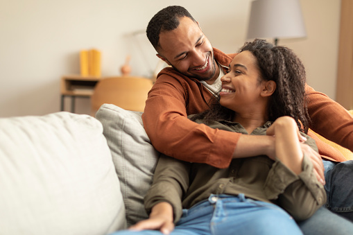 Loving black family embracing, looking at each other and smiling while relaxing on couch in living room, free space. Happy african american couple resting on sofa at home, spending weekend together