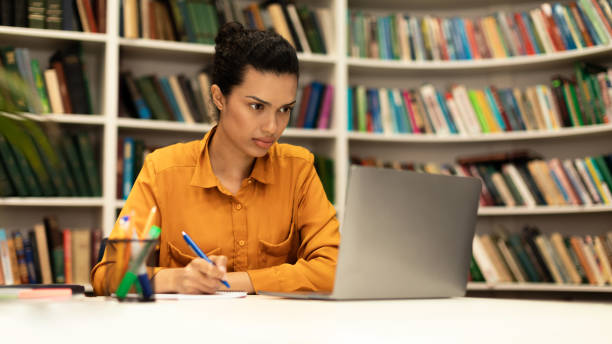 istock Focused woman using laptop and writing in notebook, studying online in library interior, panorama with free space 1471478364