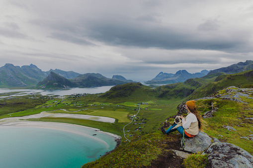 Side view of a woman and a dog on the top of the mountain, admiring the view of the turquoise-colored sea, green meadow, and dramatic peaks in Northern Norway