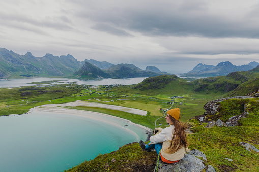 Side view of a woman and a dog on the top of the mountain, admiring the view of the turquoise-colored sea, green meadow, and dramatic peaks in Northern Norway