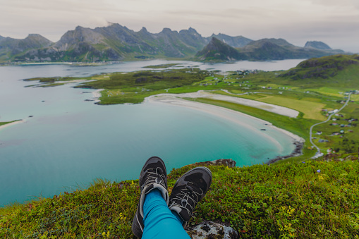 Close+up view of woman's legs relaxing on the top of the mountain, admiring the view of the turquoise-colored sea, green meadow, and dramatic peaks in Northern Norway