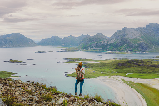 Rear view of woman traveler in a yellow hat getting to the top of the mountain, admiring the view of the turquoise-coloured sea, green meadow and dramatic peaks in Northern Norway