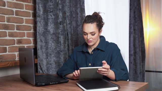 A woman works at home remotely on a computer and tablet.