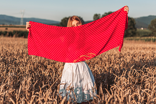 Outdoors photo of young, ginger girl in white dress holding a red scarf . Copy space