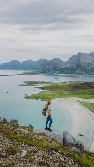 Side view of woman traveler in a yellow hat getting to the top of the mountain, admiring the view of the turquoise-coloured sea, green meadow and dramatic peaks in Northern Norway