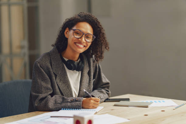istock Pretty woman freelancer making notes in notebook at desk in modern coworking and looking at camera 1471489558