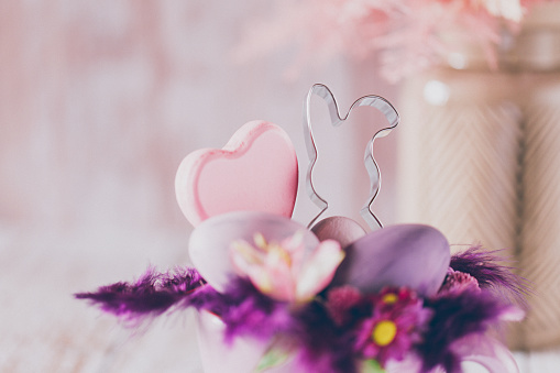 Lovely close up of a soft Easter still life of a rustic purple cup, feathers and purple eggs, a bunny cookie cutter, a heart shape in the foreground and a vase with a dried bouquet in the background. Creative color editing with added grain. Very soft and selective focus. Part of a series.