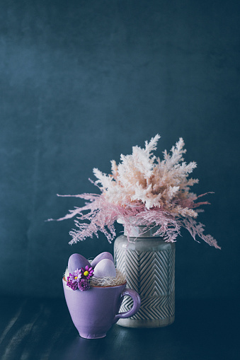 Dark low key still life of a rustic purple cup with daisies, feathers and purple Easter eggs in the foreground and a vase with dried pink and beige shrubs like pampas grass on the black background. Creative color editing with added grain. Very soft and selective focus. Part of a series.