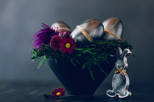 Dark low key still life of an Easter bunny and a black bowl with green moss, daisies and metallic painted Easter eggs. Creative color editing with added grain. Very soft and selective focus. Part of a series.