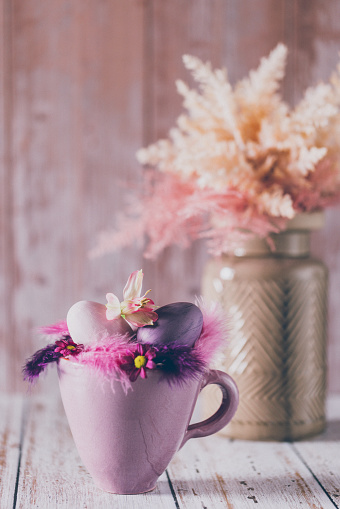 Beautiful still life of a soft Easter still life of a rustic purple cup, feathers and purple eggs in the foreground and a vase with dried pink and beige shrubs like boho pampas grass on a salmon pink wooden background. Creative color editing with added grain. Very soft and selective focus. Part of a series.