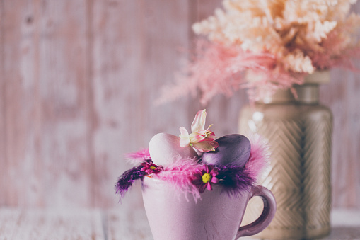 Beautiful close up of a soft Easter still life of a rustic purple cup, feathers and purple eggs in the foreground and a vase with dried pink and beige shrubs like boho pampas grass on a salmon pink wooden background. Creative color editing with added grain. Very soft and selective focus. Part of a series.