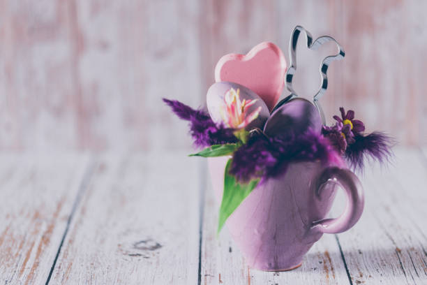 Still life of a rustic purple cup with an Easter bunny cookie cutter, daisies, feathers a heart shape and purple Easter eggs on wooden background with copy space. Creative color editing with added grain. Very soft and selective focus. Part of a series.
