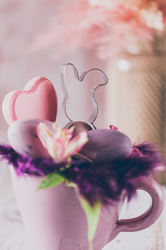 Beautiful close up of a soft Easter still life of a rustic purple cup, feathers and purple eggs, a bunny cookie cutter, a heart shape in the foreground and a vase with dried pink and beige shrubs like boho pampas grass on a salmon pink wooden background. Creative color editing with added grain. Very soft and selective focus. Part of a series.