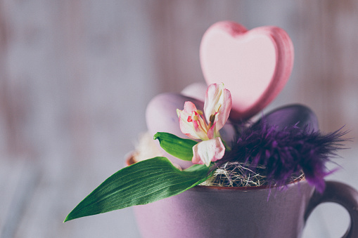 Close up of a rustic purple cup with an Easter eggs, a spring flower, feathers a heart shape on unfocused wooden background with copy space. Creative color editing with added grain. Very soft and selective focus. Part of a series.