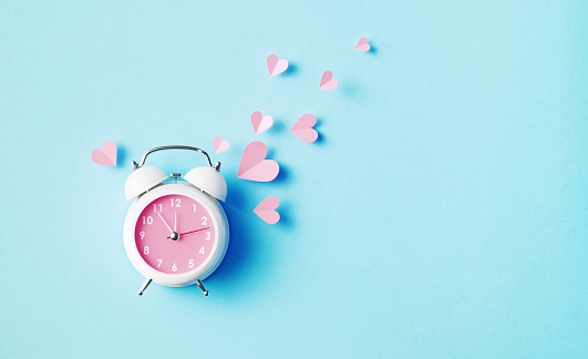 Pink hearts scattered from a white alarm clock on blue background. Horizontal composition with copy space.
