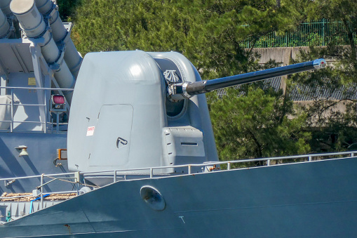 The forward turret and 5 inch Mk45 Mod 2 automatic rapid fire gun of HMAS Arunta, a frigate of the Royal Australian Navy.  Also visible are the canisters of her anti-ship missiles.  A red kangaroo insignia is visible on the cap covering the gun barrel.  She is docked at Garden Island, Sydney Harbour in preparation for an open day to the public during Navy Week, the first open day since the Covid pandemic.  This image was taken on a sunny afternoon from Woolloomooloo Bay on 25 February 2023.