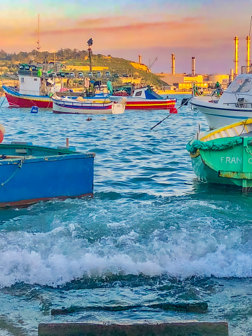 Marsaxlokk, Malta - November, 2018: Waves and boats in the harbour of Marsaxlokk during sunset, picture taken from the waterfront.