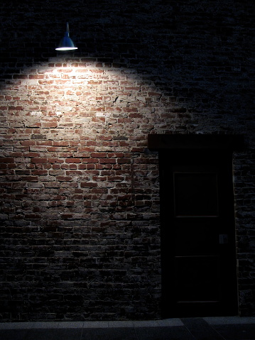 A single flood light partially lights up a red brick wall and a shaded dark door. Taken in an alley in San Francisco, California, USA.