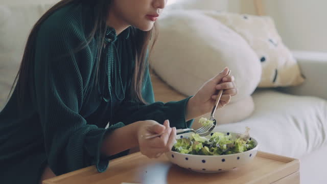 Young happy asian woman eating healthy salad sitting on the sofa in living room.