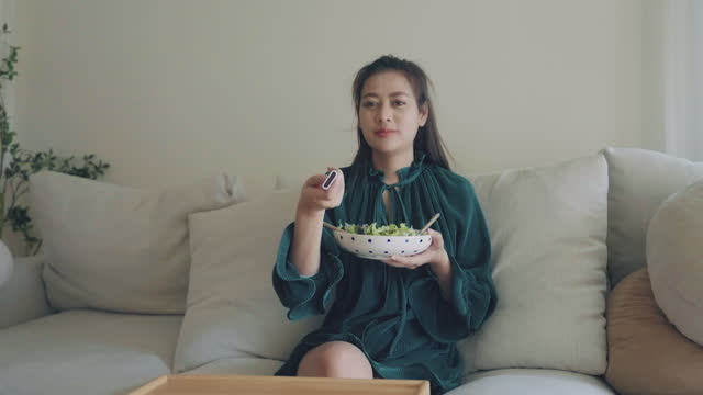 Young happy asian woman eating healthy salad sitting on the sofa in living room.