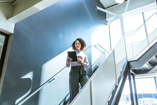 Biracial woman walking down the stairs of a modern office, holding a digital tablet