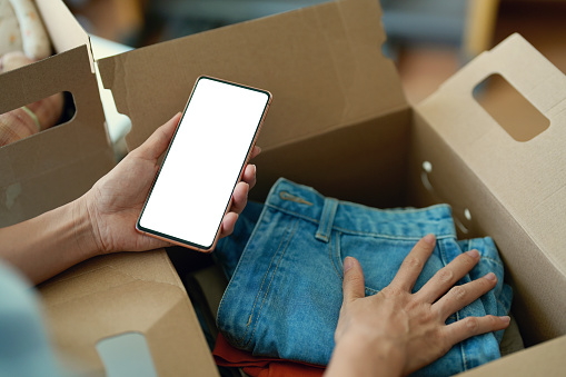 Cropped image of an Asian female woman using a blank screen smartphone while packing or unpacking as she is moving into a new place house. she is using her smartphone chatting with friends and family or finding a delivery service nearby.