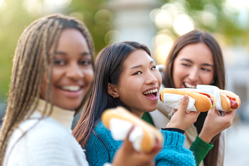 Selective focus on an asian woman eating hot dog with friends in a park