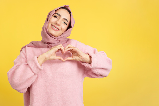 Muslim woman representing a heart shape of fingers in studio with yellow background