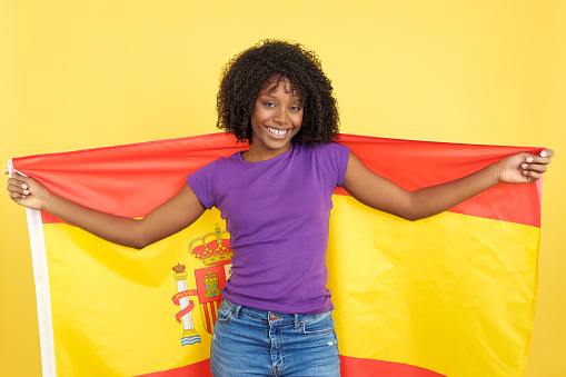 Happy afro woman smiling and raising a spanish national flag in studio with yellow background