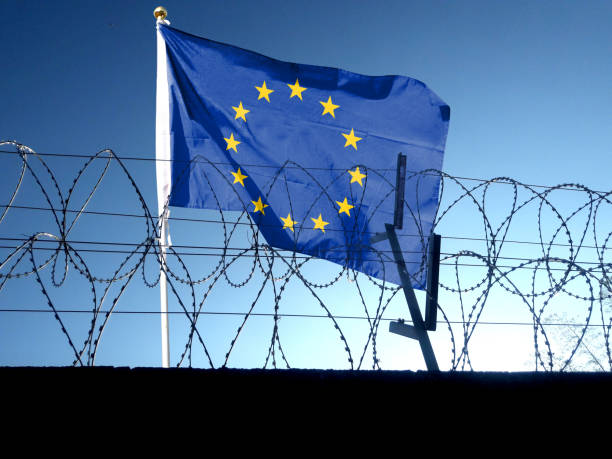 European Union flag waving behind barbed wire fence under a clear blue sky.