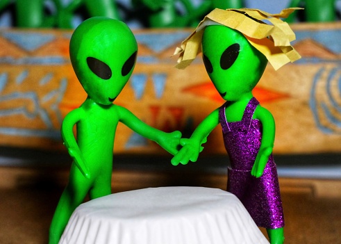 Two green aliens on a date.  One just great and the other with blonde hair and in a purple sparkling dress.  Large black eyes.  Photo includes a white table.