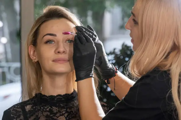 Makeup artist in black gloves colors eyebrows to client neatly. Professional eyebrows tinting in modern beauty salon closeup on blurred background