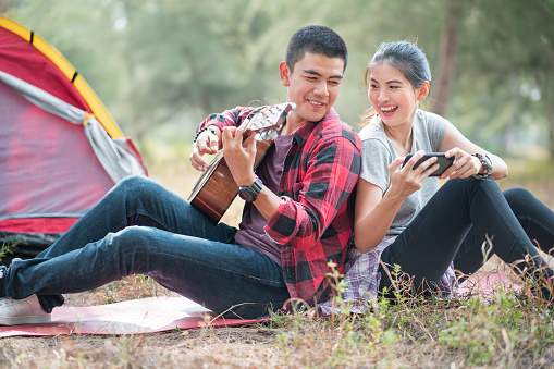 Romantic couple camping outdoors and sitting at tent. Happy couple on a romantic camping vacation. Couple asian camp forest adventure and playing guitar together in nature. Travel in summer holiday
