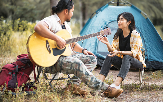 Romantic couple camping outdoors and sitting at tent. Happy couple on a romantic camping vacation. Couple asian camp forest adventure and playing guitar together in nature. Travel in summer holiday