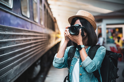 Tourist woman taking a photo with camera of side of train on railway background. Young asian woman traveler with backpack in the railway, Travel concept