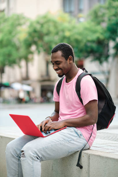 istock Man with backpack smiling while using a laptop sitting on a bench outdoors. 1471583535