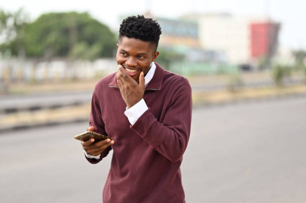 Attractive young african american man on the street holding his smartphone, smiling happy laughing expression demonstrating with his hands to his jaw