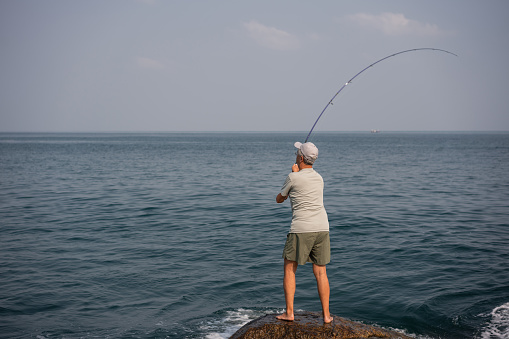 Senior fisherman casting out his fishing line into the sea from the rocky shoreline.
